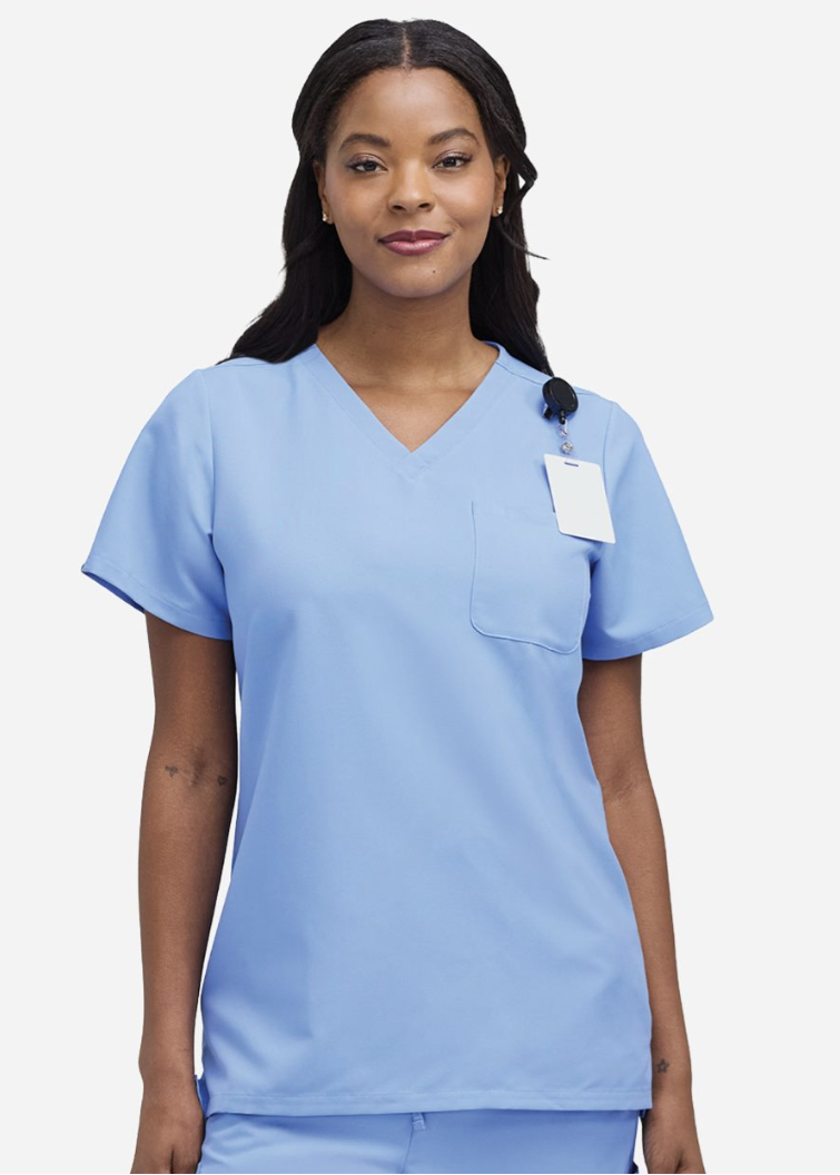 A woman modeling a light blue V-neck scrub top with a chest pocket and a blank white hangtag, set against a plain light gray background.